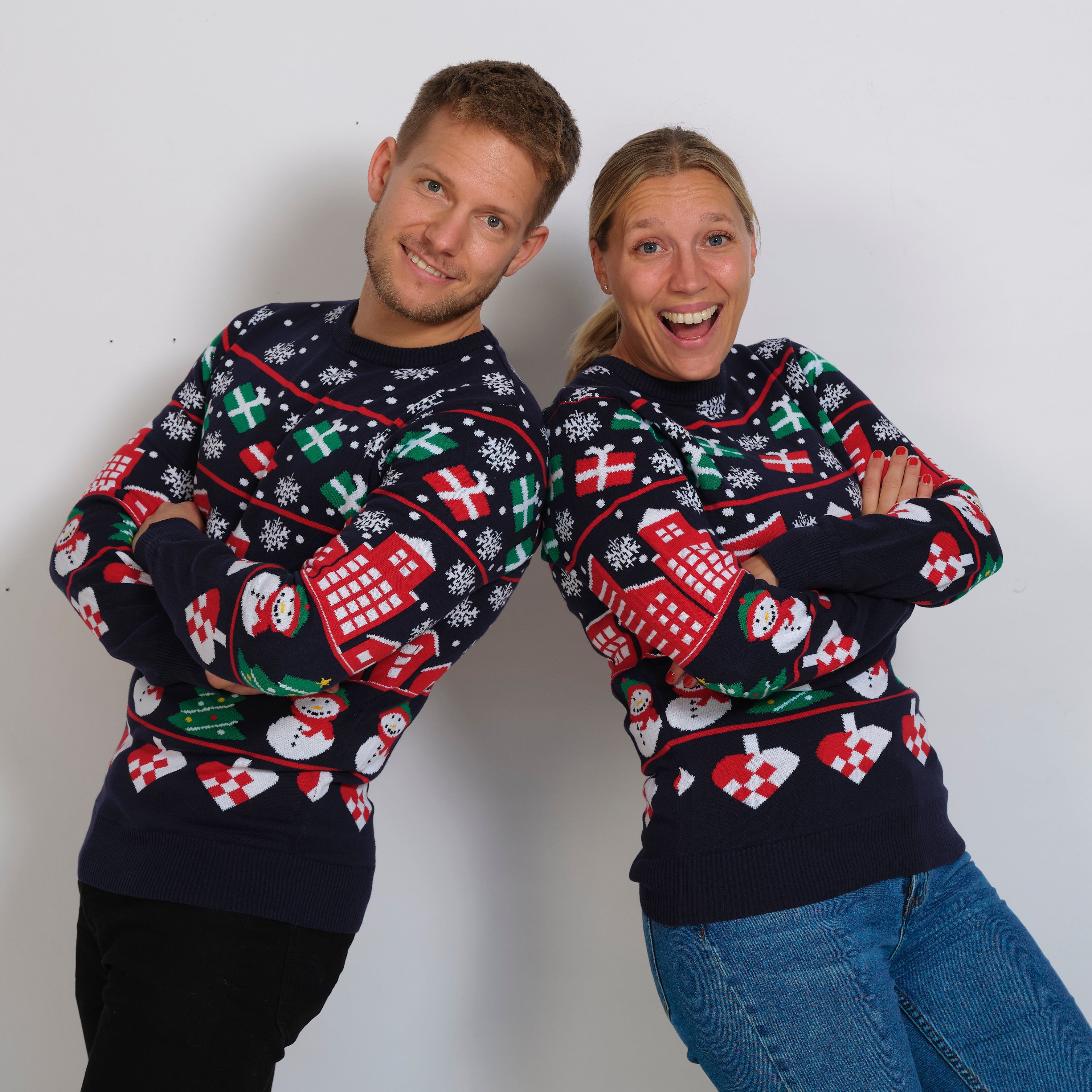 En mand og en dame, der smiler, læner sig op ad hinanden, og som begge har en julesweater på.